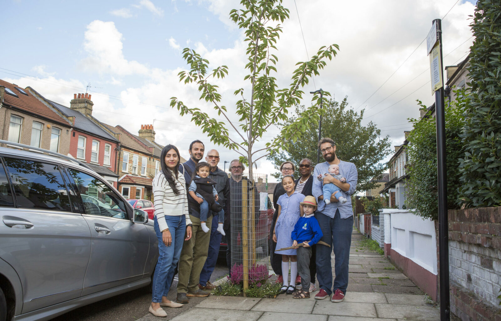 A group of neighbours standing around their newly sponsored tree on a sunny blue sky day